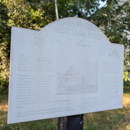 A faded Heritage Home sign for the Altnow-Smith Home beside trees and a white fence